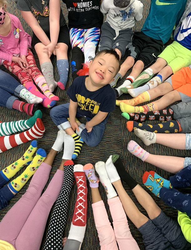 At Clarks Creek, Kindergarten students surround their friend with their brightly-color socks as part of the #LotsOfSocks celebration